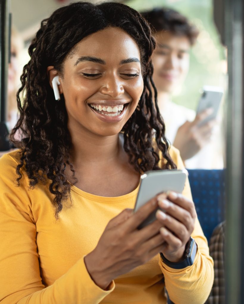 Happy black lady using smartphone wearing headphones in bus